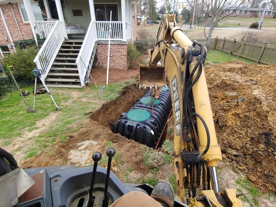 View from backhoe cab of a black plastic septic tank with green lids set in a trench beside a brick house