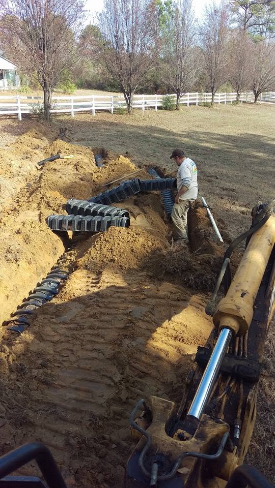 Worker placing black plastic infiltrator chambers in excavated septic trenches beside a backhoe on a sloped lot.
