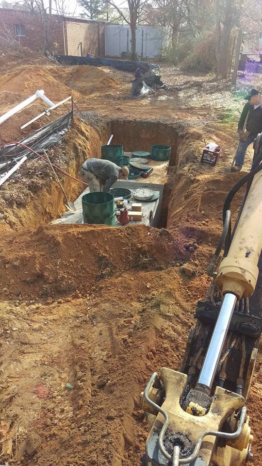 Two workers fit green risers onto concrete septic tanks in an excavated pit, with a backhoe boom in the foreground.