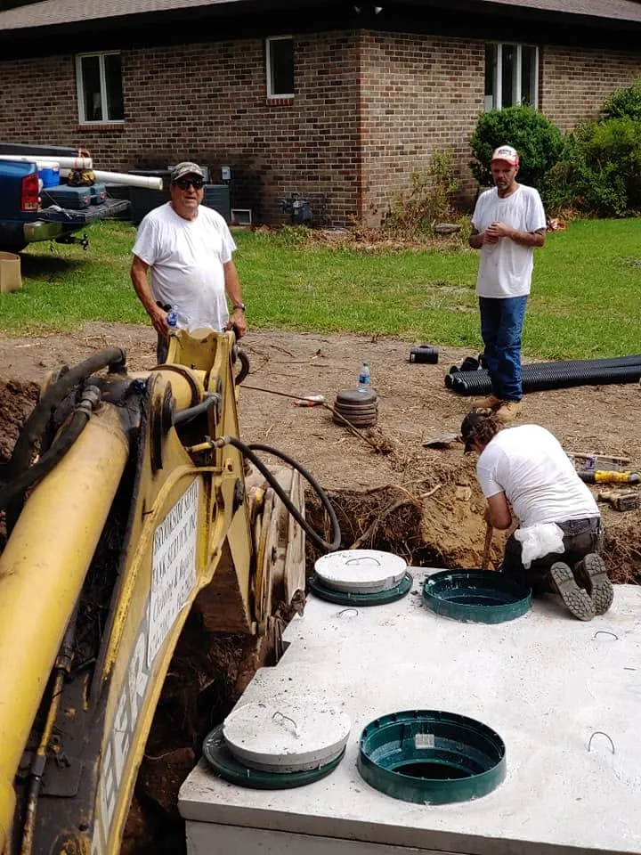 Three workers install a concrete septic tank with green riser lids beside a brick home while a backhoe sits nearby.