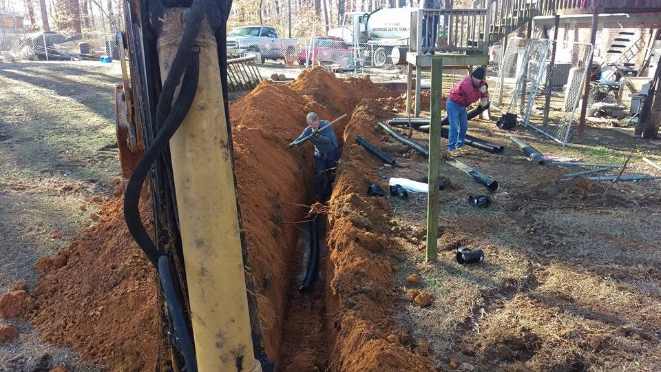 Two workers install black corrugated septic pipe in a red clay trench beside a backhoe in a residential backyard.