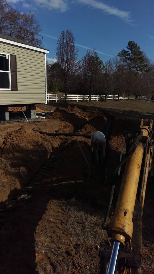 View from a backhoe over excavated trenches with corrugated drain pipe beside a manufactured home and white fence.