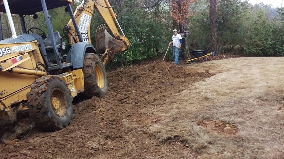 Yellow backhoe grading dirt in a wooded backyard while a worker rakes soil beside a wheelbarrow