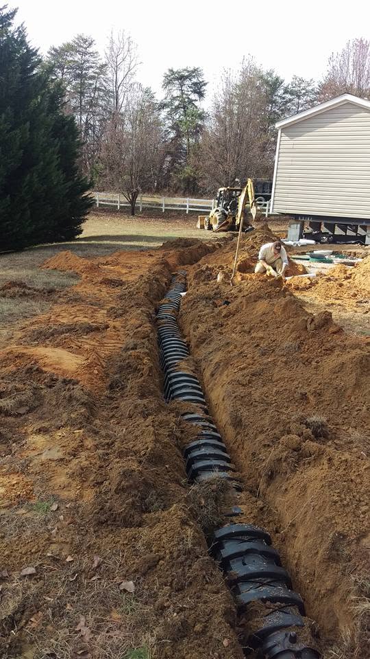 Long trench with black plastic drain field chambers beside a mobile home, with a worker and backhoe in the background