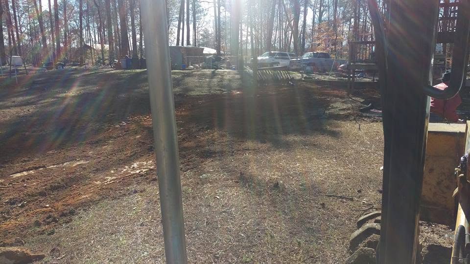 View from excavator cab of a freshly graded and backfilled yard on a wooded residential lot with trucks in the background.