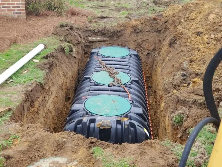 Black plastic septic tank with three green riser lids and a lifting chain set in an excavated pit beside a brick home.