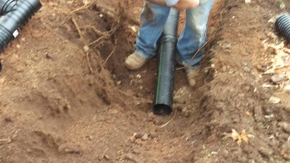 Worker in jeans standing in a dirt trench positioning a black corrugated drain pipe into freshly dug soil