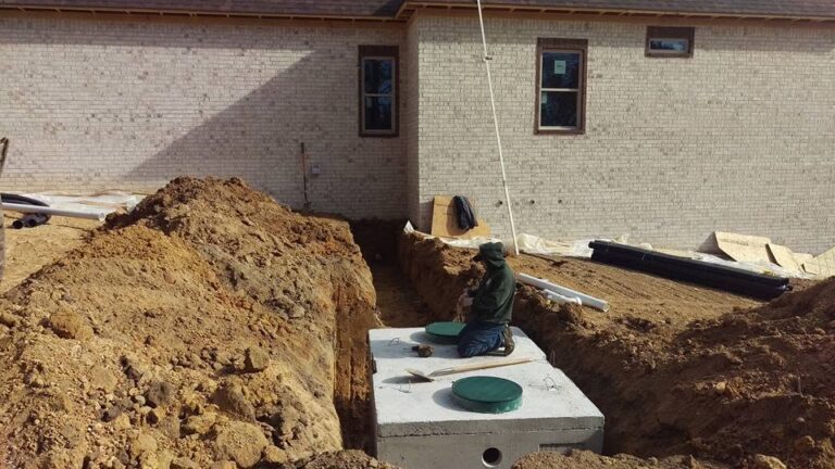 Worker kneels on a concrete septic tank with green riser lids in an excavated pit beside a new brick house.