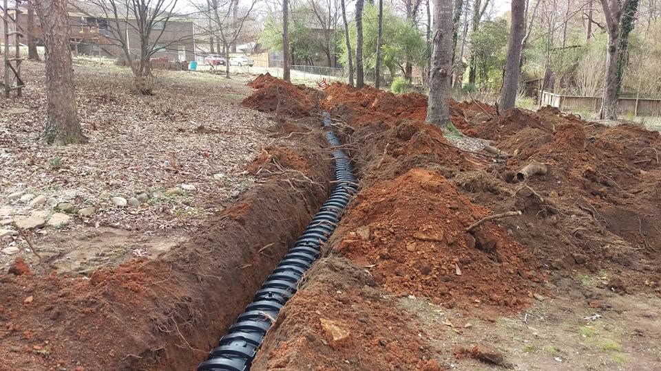 Long open trench with black corrugated septic pipe running through a wooded residential yard with red clay soil.
