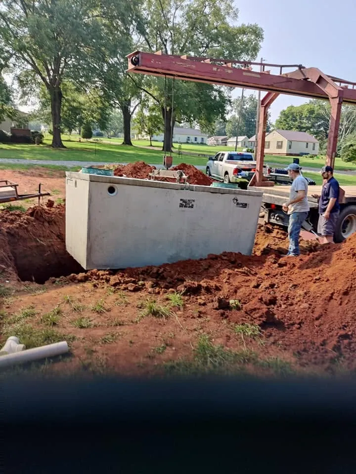 Two workers guide a precast concrete septic tank lowered by truck crane into a red clay excavation in a residential yard