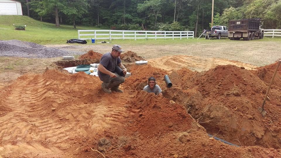 Two workers in a red clay excavation connect black pipe near a buried septic tank by a white fenced pasture with a dump truck
