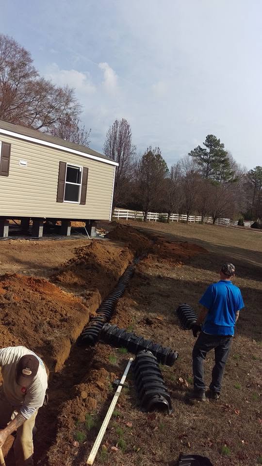 Two workers installing black plastic septic chambers in trenches beside a mobile home on a rural Alabama lot.