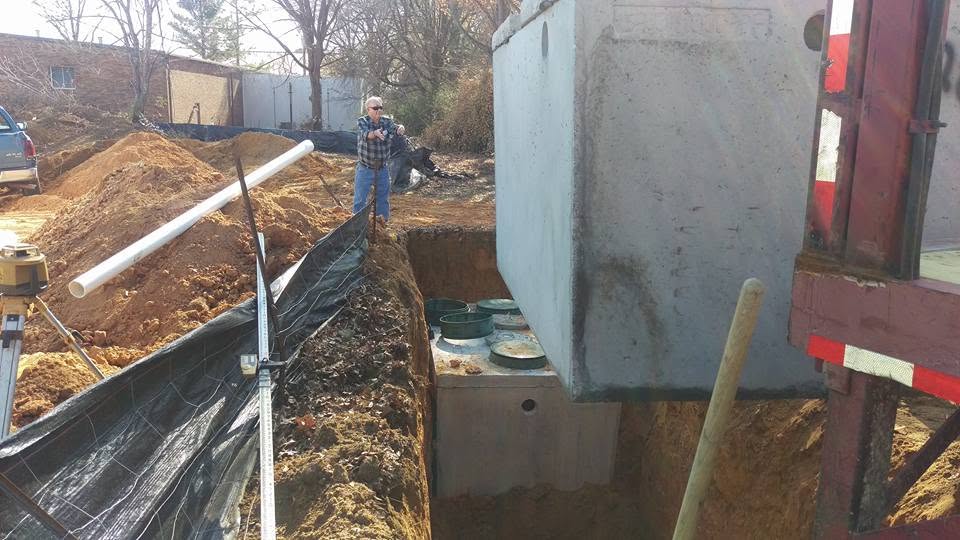 Concrete septic tank lid lowered from a delivery truck over an open excavation while a worker watches, Birmingham AL