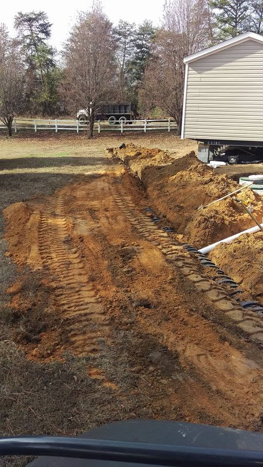 Long excavated trench with black septic chambers installed beside a manufactured home with a white fence in the background.