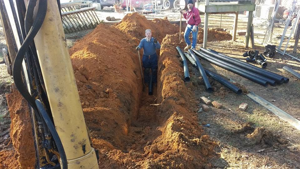 Two workers in a deep red clay septic trench beside stacked corrugated drain pipe, seen past an excavator boom.