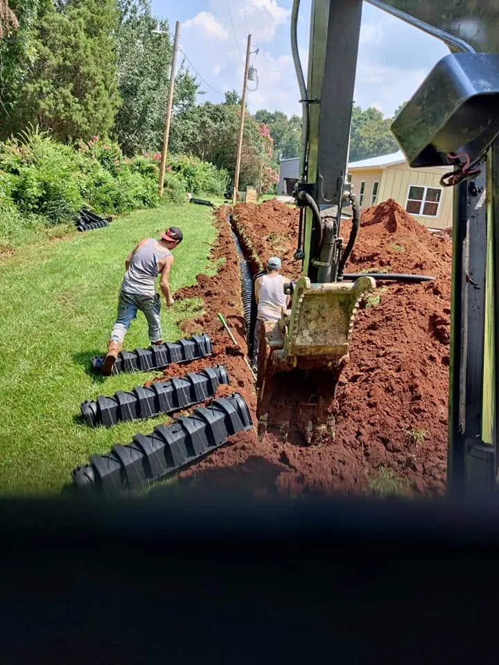 View from an excavator cab of two workers installing black drain field chambers in a red clay trench beside a house.