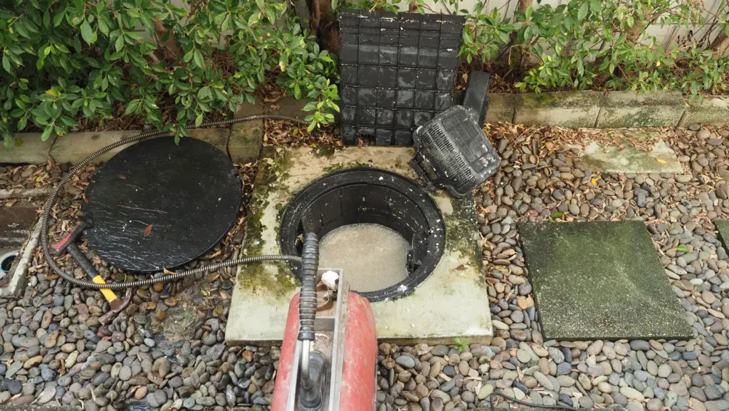 Open in-ground grease trap with lid and strainer basket removed, drain snake cable feeding into the tank beside a red
