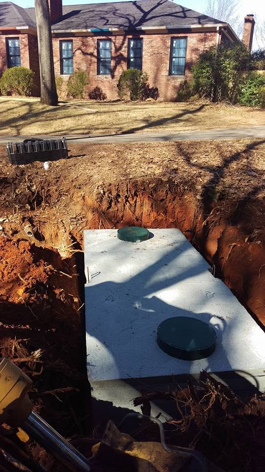 Concrete septic tank with two green riser lids set in an excavated pit in front of a brick house