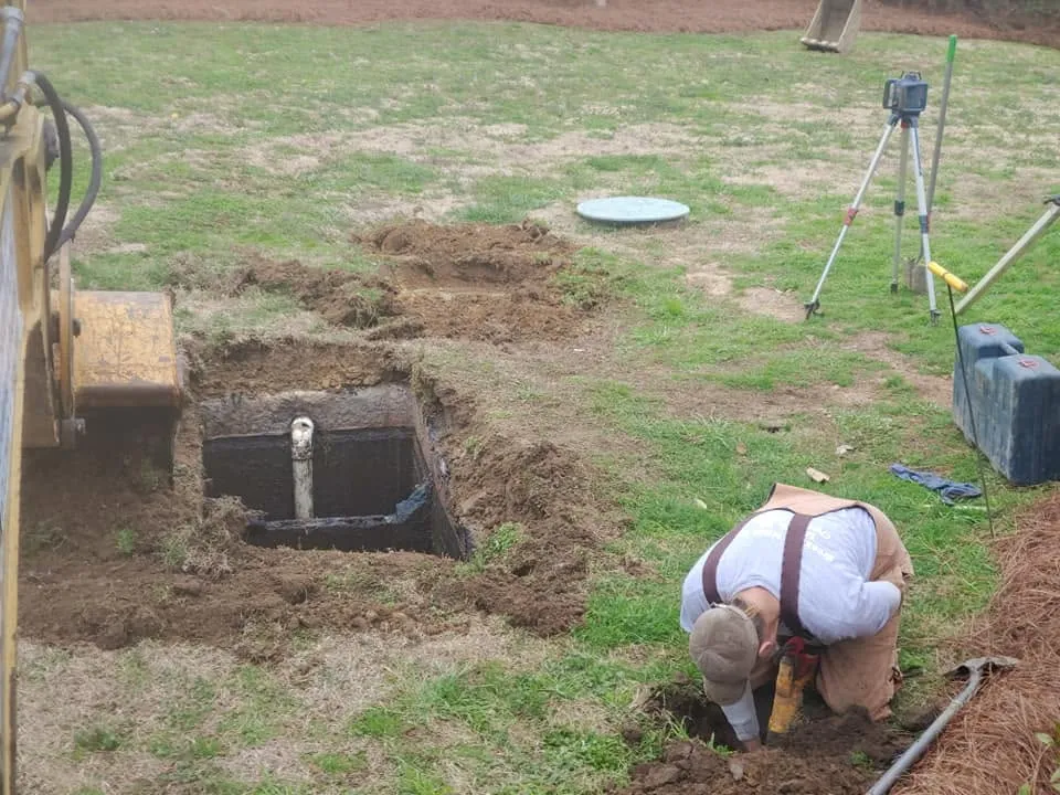 Worker kneels digging by hand near an opened septic tank pit with a white pipe, laser level on tripod nearby in a grassy yard