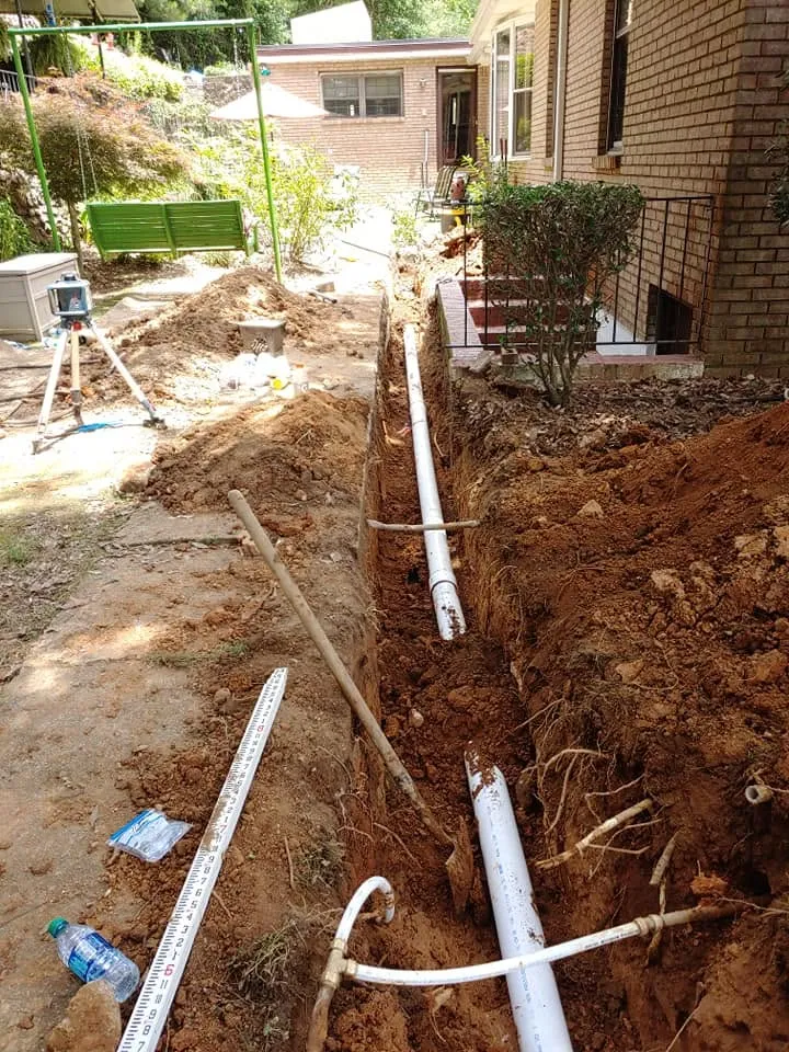 White PVC pipe laid in a dirt trench beside a brick house, with a laser level tripod and grade rod nearby