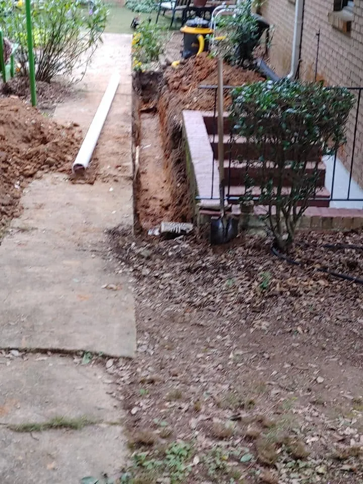 Narrow hand-dug trench in red clay along a concrete walkway beside brick steps, with a white PVC pipe and shovel nearby