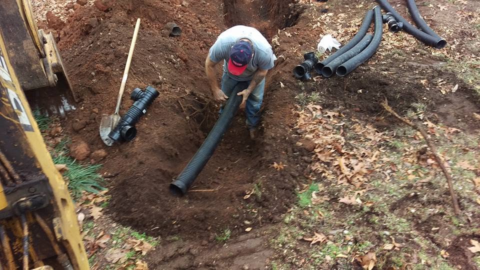 Worker in a red clay trench holding a length of black corrugated drain pipe, with fittings and a backhoe nearby.