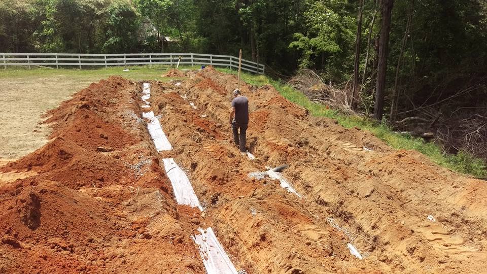 Worker walks between parallel gravel-lined drain field trenches dug in red clay on a fenced rural lot