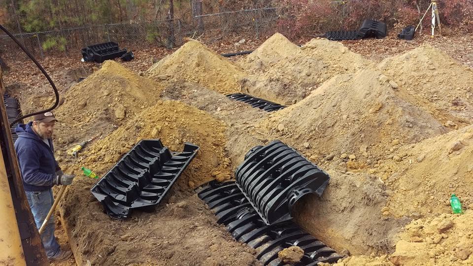 Worker with a shovel beside parallel trenches holding black plastic septic chambers on an excavated wooded lot.