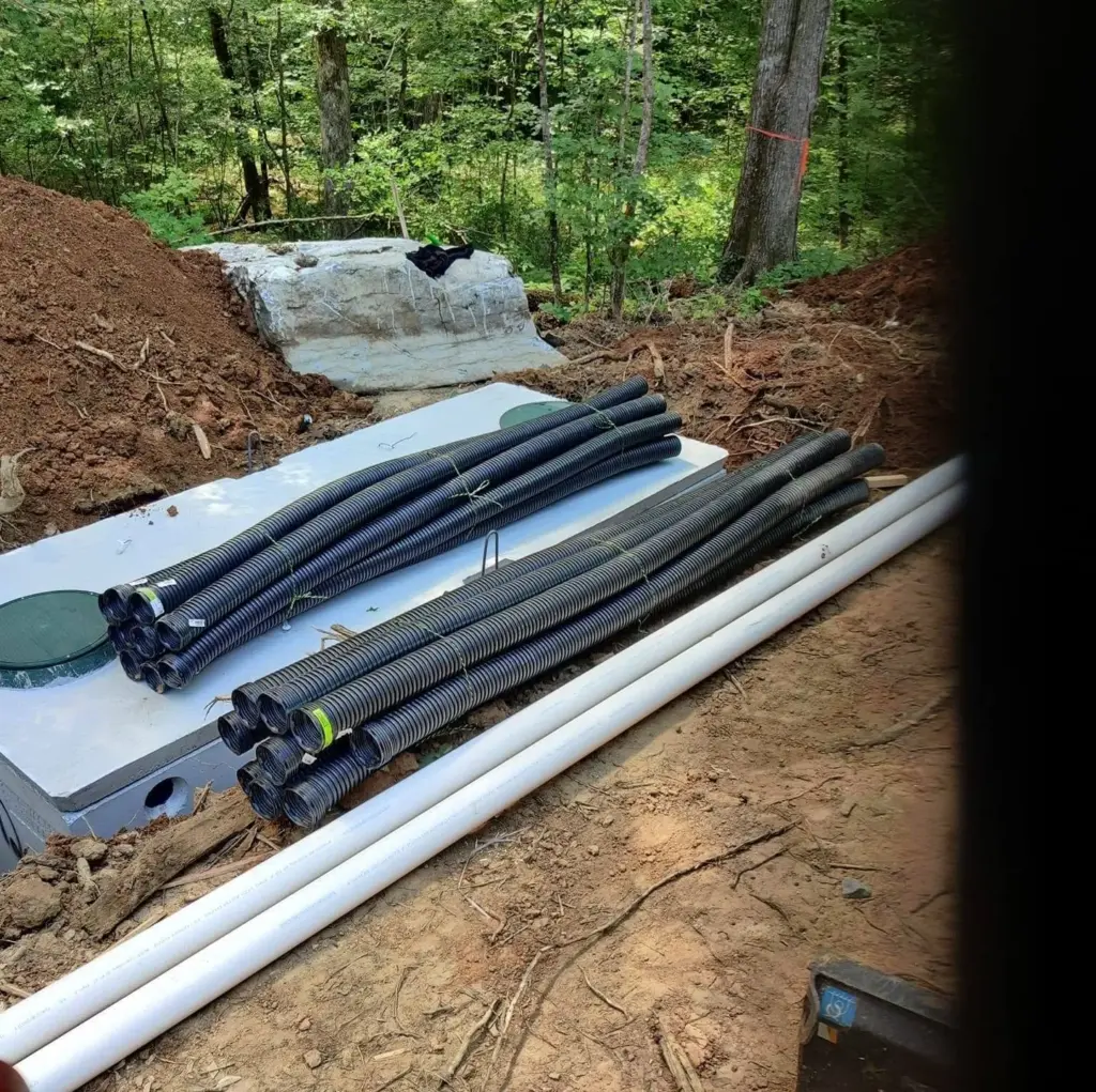 Bundles of black corrugated drain pipe and white PVC pipe staged on a concrete septic tank lid at a wooded job site.