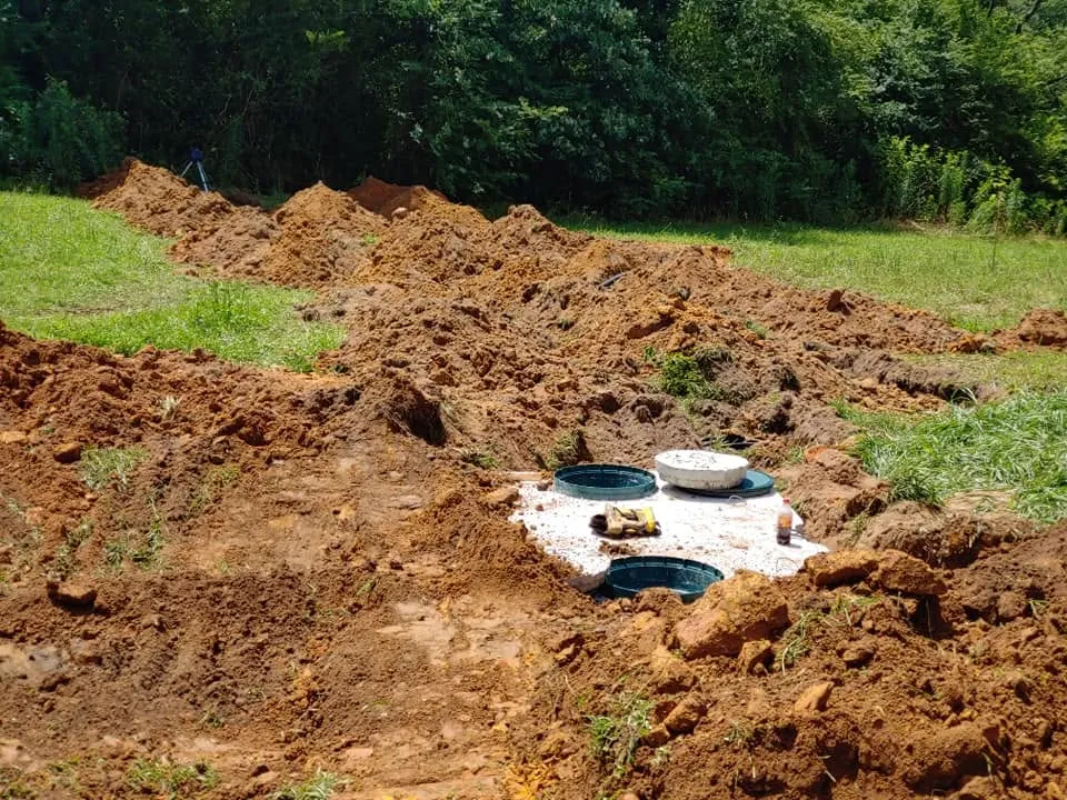 Top of a buried concrete septic tank with green risers surrounded by mounds of red clay soil in a grassy yard.