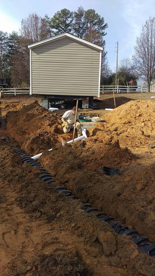 Worker kneels connecting white PVC pipe to a black septic chamber trench in front of a mobile home on a dirt lot.