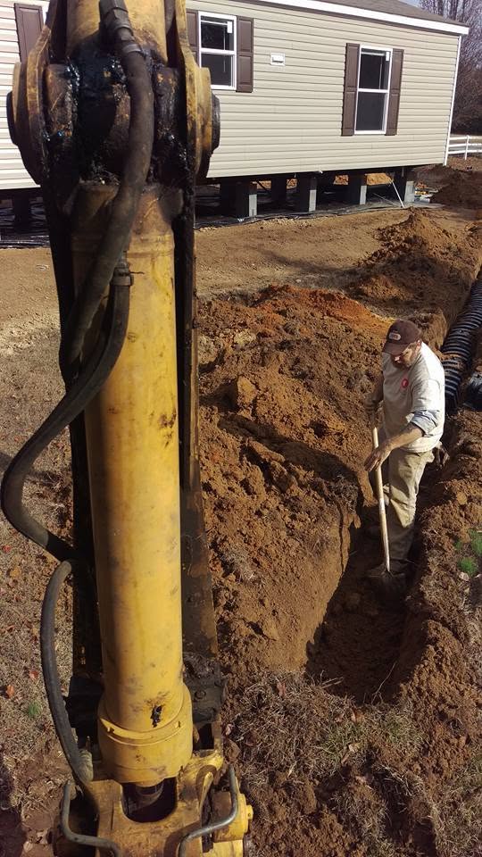 Worker with shovel digging a trench beside an excavator arm and corrugated pipe near a mobile home