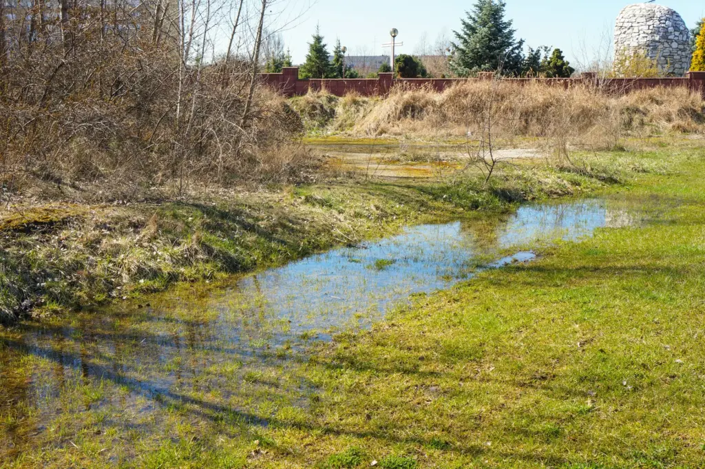Standing water pooled across a grassy lawn beside dry brush and a red fence, with a stone structure in the background