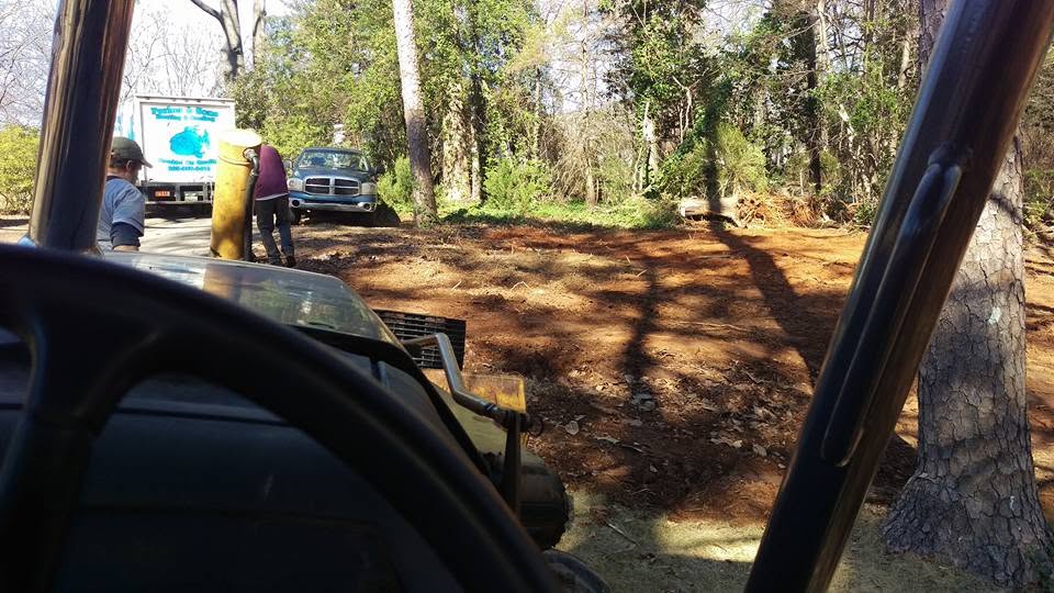 View from tractor cab over cleared red clay ground toward workers and trucks at a wooded septic job site
