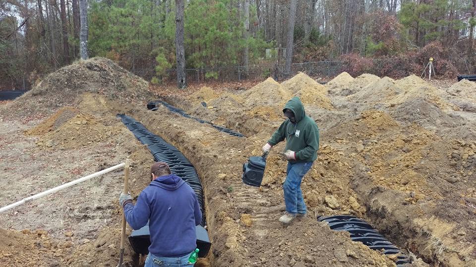 Two workers placing black plastic drain field chambers in dug trenches on a wooded lot with soil piles