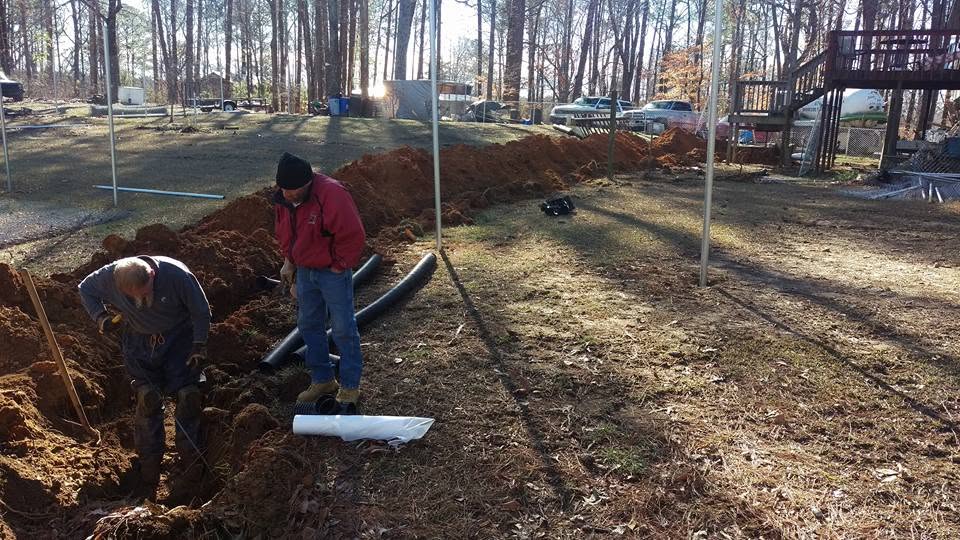 Two workers dig a septic drain line trench in a backyard with corrugated black pipe and red clay soil piled alongside