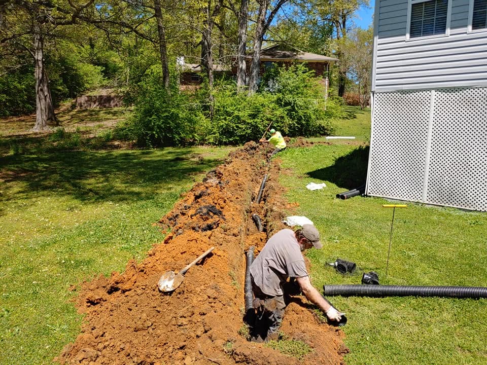 Two workers install corrugated drain pipe in a long red clay trench across a grassy backyard beside a house