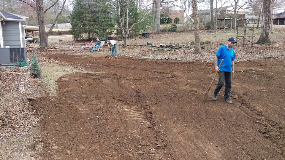 Two workers with shovels grading fresh red dirt over a backfilled septic system in a residential backyard