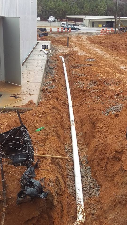 White PVC sewer pipe laid on a gravel bed in a long trench beside a metal commercial building in red clay soil.