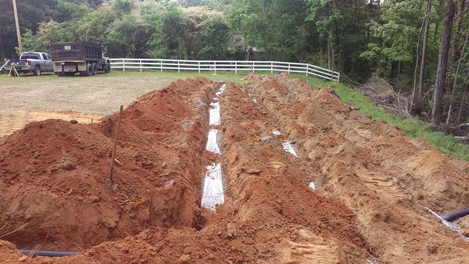Rows of excavated drain field trenches in red clay soil with a dump truck and white fence in the background