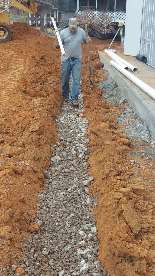 Worker carrying PVC pipe and shovel walks along a gravel-lined septic drain field trench cut into red clay soil.