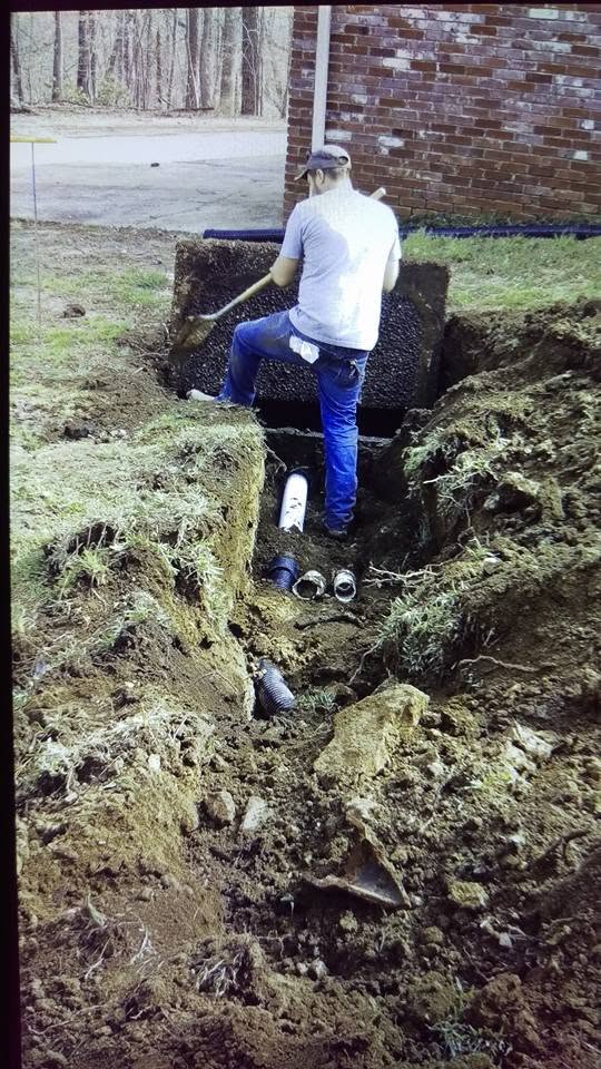 Worker with a shovel digs a septic trench beside a brick house, with corrugated pipe fittings on the ground nearby.