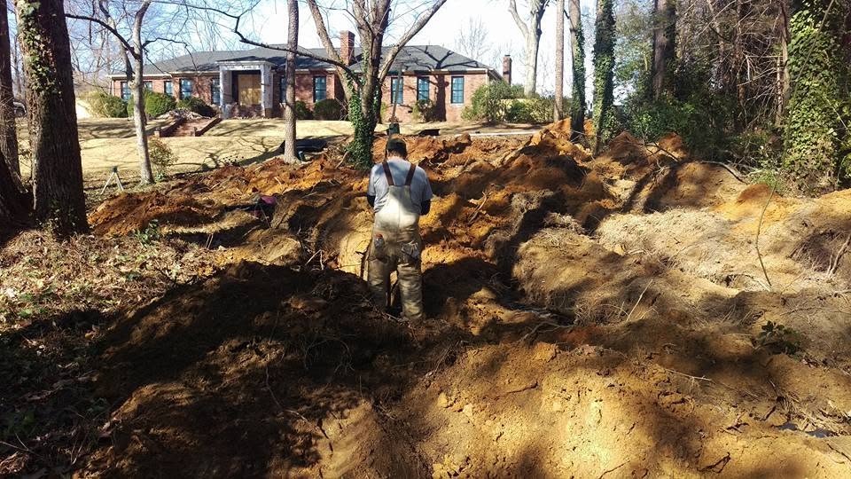 Worker in overalls standing in excavated septic trenches in the wooded front yard of a brick house.