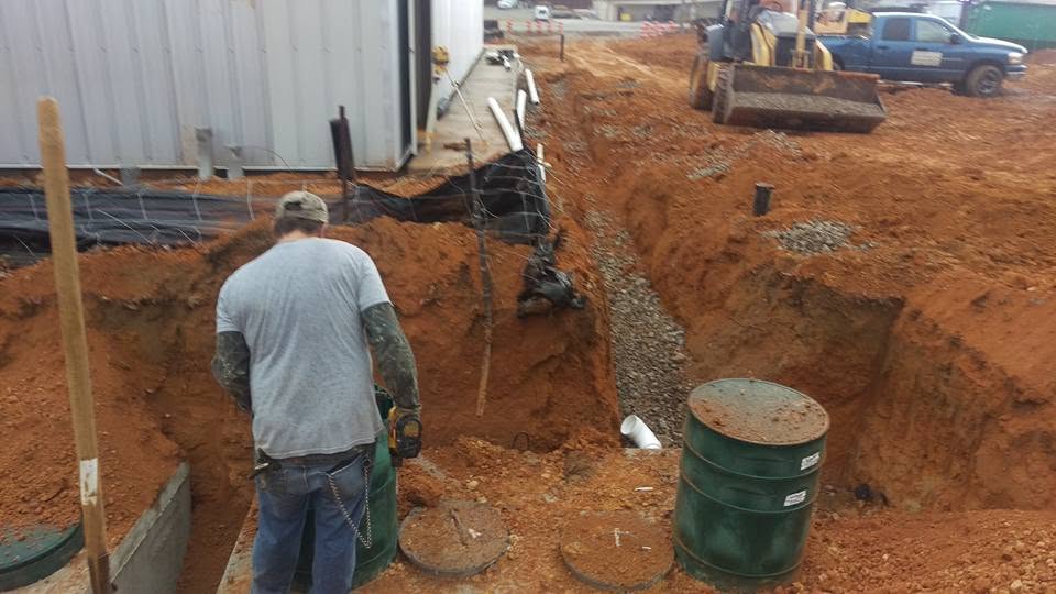 Worker beside gravel-lined septic trench with green risers and buried concrete tank lids at a commercial site in red clay