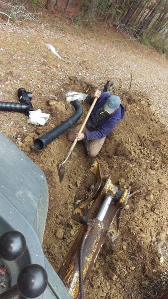 Worker kneeling in dirt with a shovel connecting black corrugated septic pipe beside a backhoe boom on a wooded lot