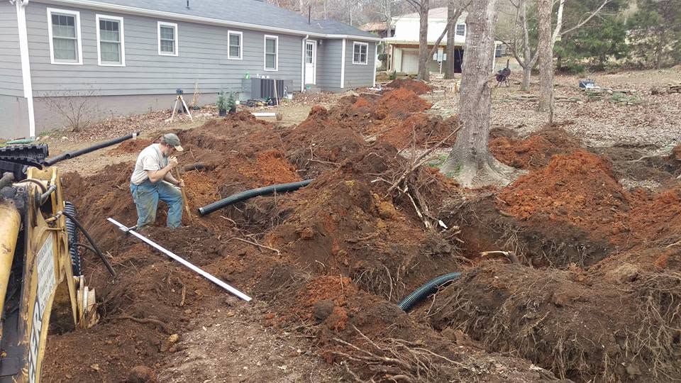 Worker in an open trench installing black corrugated septic field line pipe beside a gray house with a backhoe nearby.