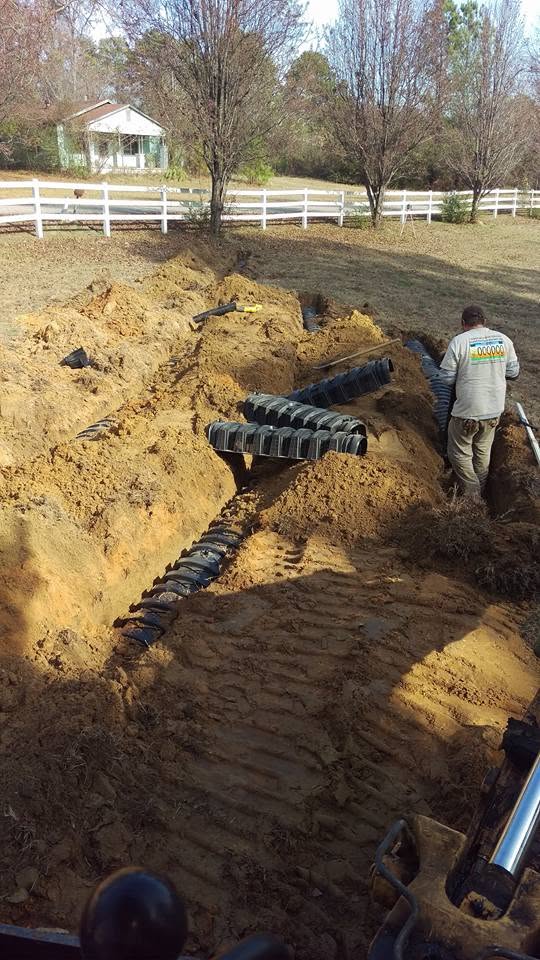 Worker placing plastic infiltrator chambers in excavated drain field trenches at a rural home with a white rail fence.