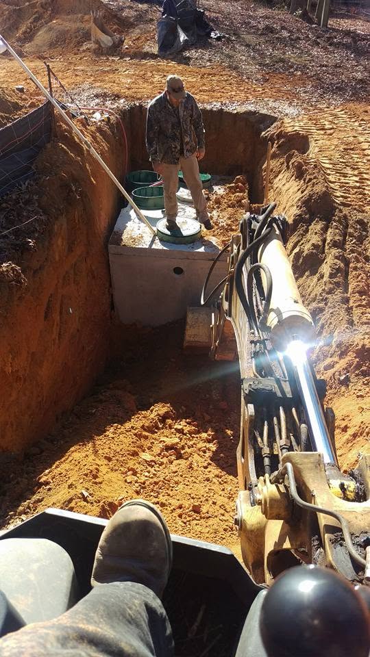 Worker stands on a concrete septic tank with green risers inside a deep red clay excavation, viewed from a backhoe seat.