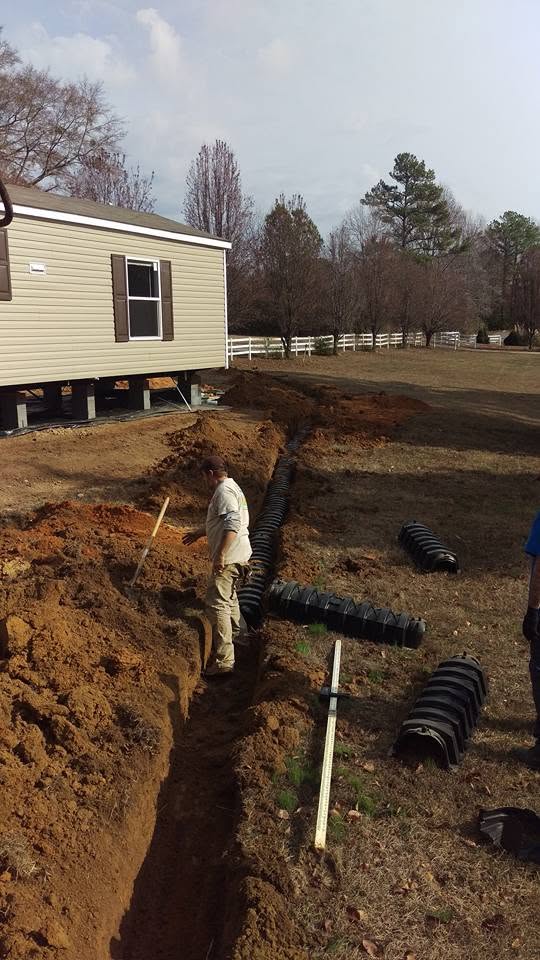 Worker stands in a dirt trench beside a mobile home with black septic chambers staged on the ground nearby