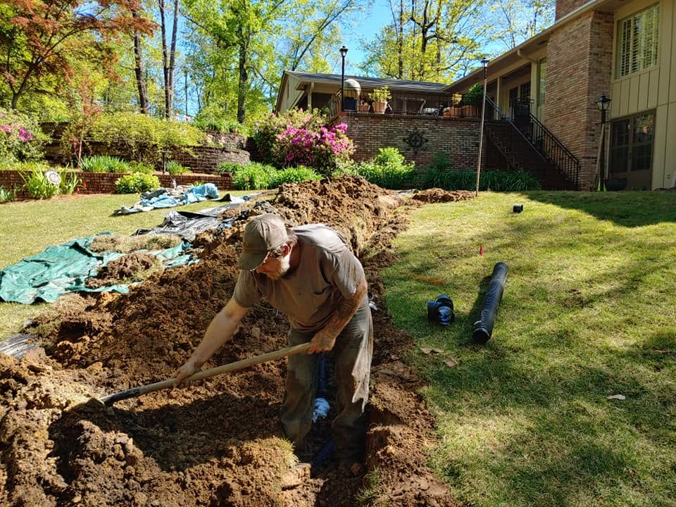 Worker shoveling soil in an open drain field trench behind a brick home with corrugated pipe on the lawn